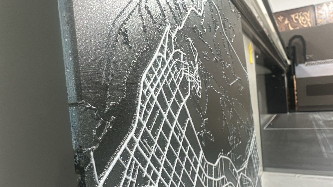 Close-up of a city street map in raised white lines on a dark panel, giving an etched look, with a flatbed printer behind