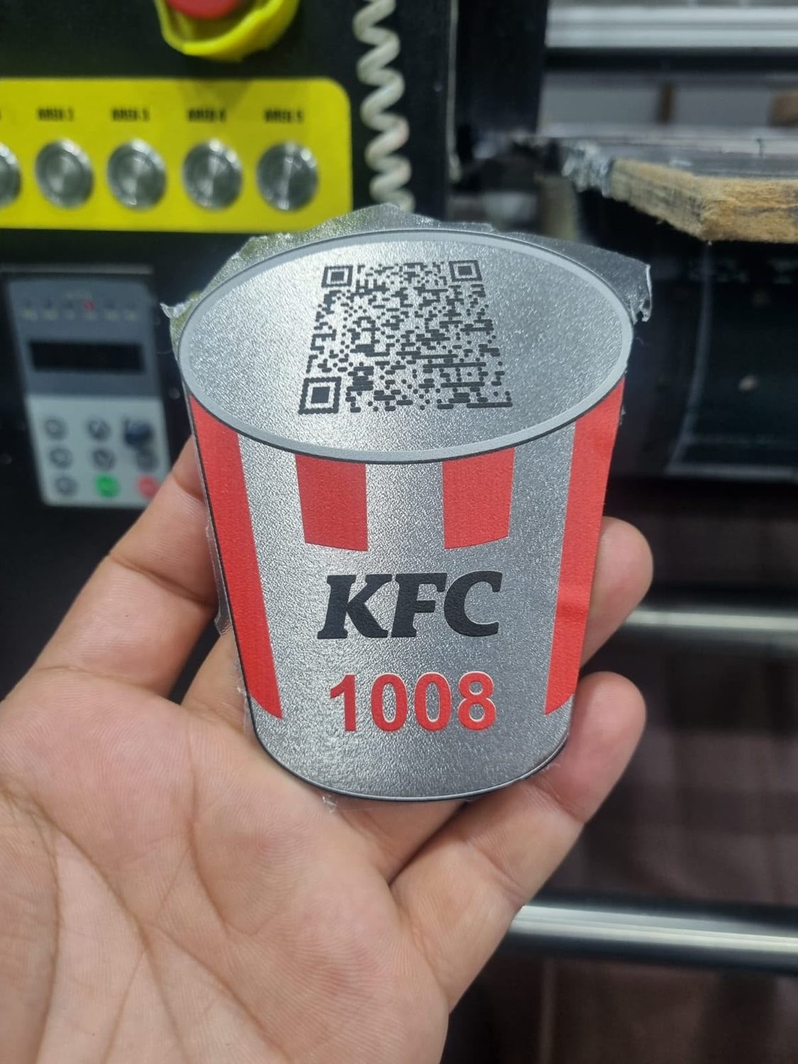 A die-cut metal tag printed in full colour with a QR code on stippled silver stock, held in the workshop